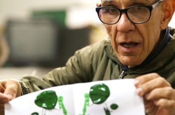 Foto de um senhor grisalho, com óculos, segurando uma folha com pintura abstrata em tinta verde