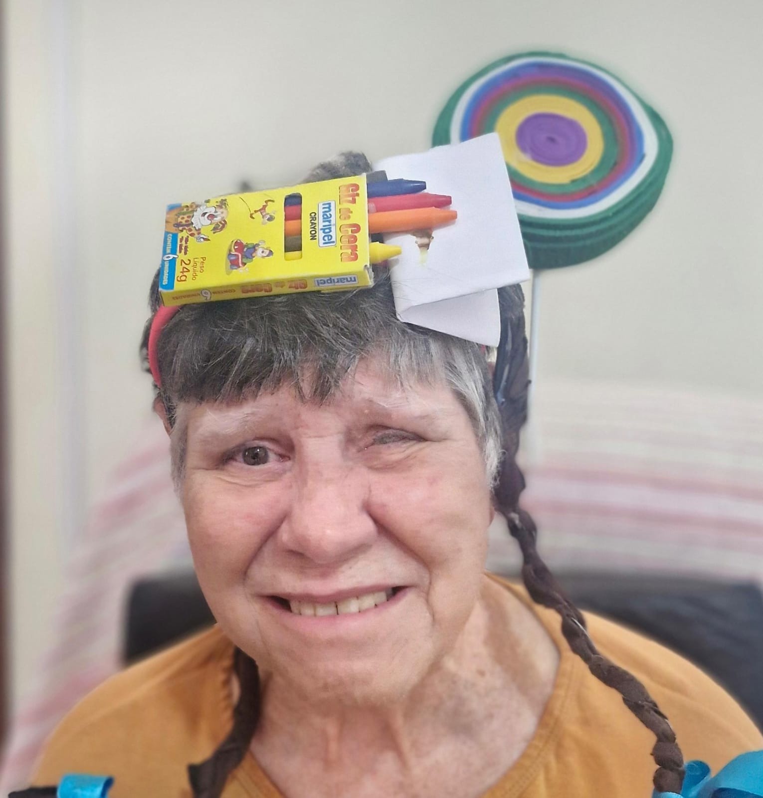 Uma mulher idosa sorri usando um enfeite criativo na cabeça, feito com lápis de cor em uma caixa e detalhes coloridos ao redor