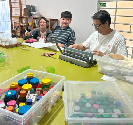 Três pessoas adultas estão sentadas ao redor de uma mesa de atividades, sorrindo enquanto trabalham com materiais coloridos. Há potes de tinta e tampinhas organizadas em caixas transparentes sobre a mesa.