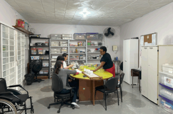 Um grupo de quatro pessoas está sentado ao redor de uma mesa grande em uma sala organizada com prateleiras cheias de materiais e caixas etiquetadas. Uma profissional orienta a atividade.