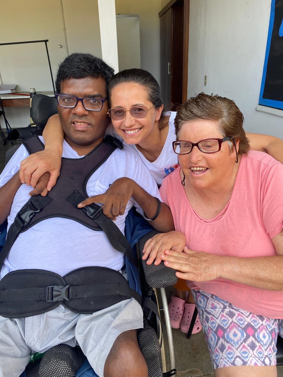 Uma mulher abraça duas pessoas adultas, uma delas em cadeira de rodas. Todos estão sorrindo em um momento de carinho ao ar livre